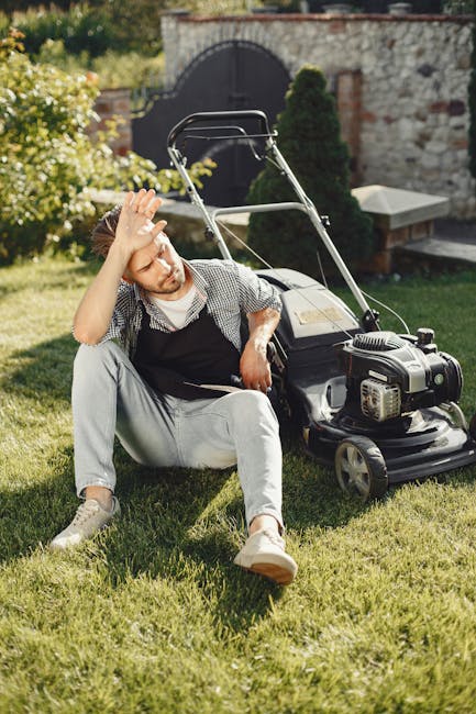 A young man sitting on a well-maintained lawn appears to be experiencing frustration or fatigue, with one hand resting on his forehead. He is wearing light-colored pants, a checkered shirt, and an apr
